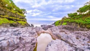 Rocks with a pool in the middle surrounded by green covered cliffs opening out to the ocean beyond